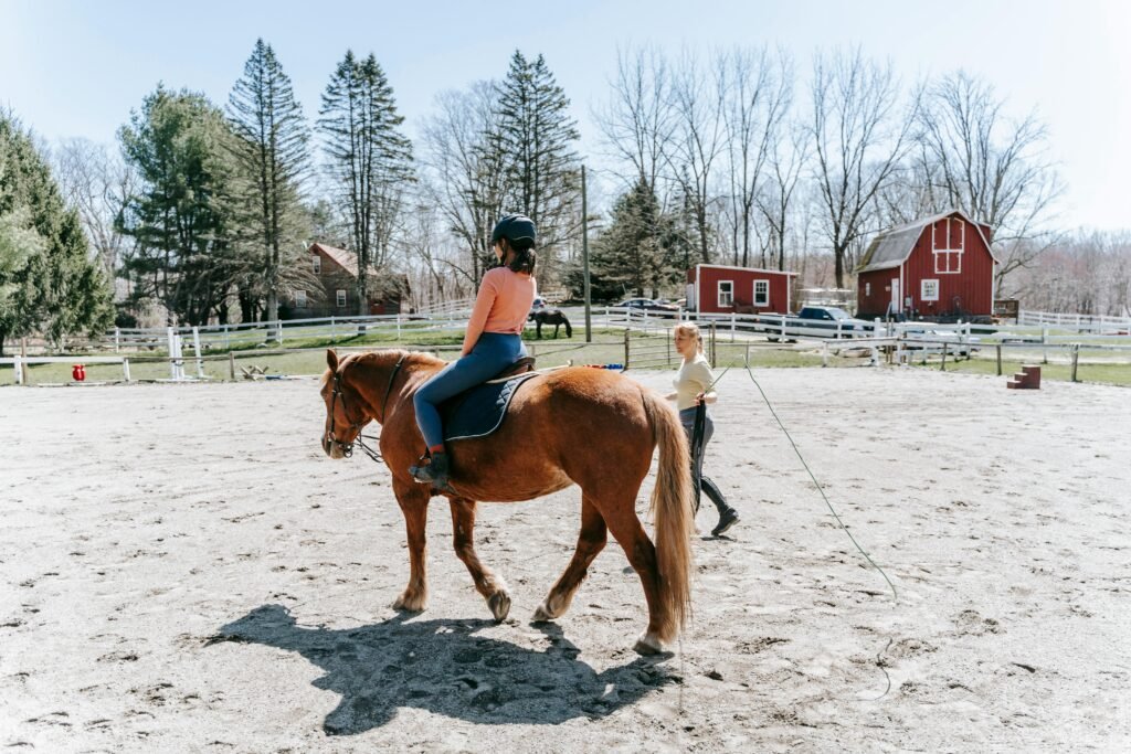 A woman learning horseback riding in an outdoor arena with instructor guidance.